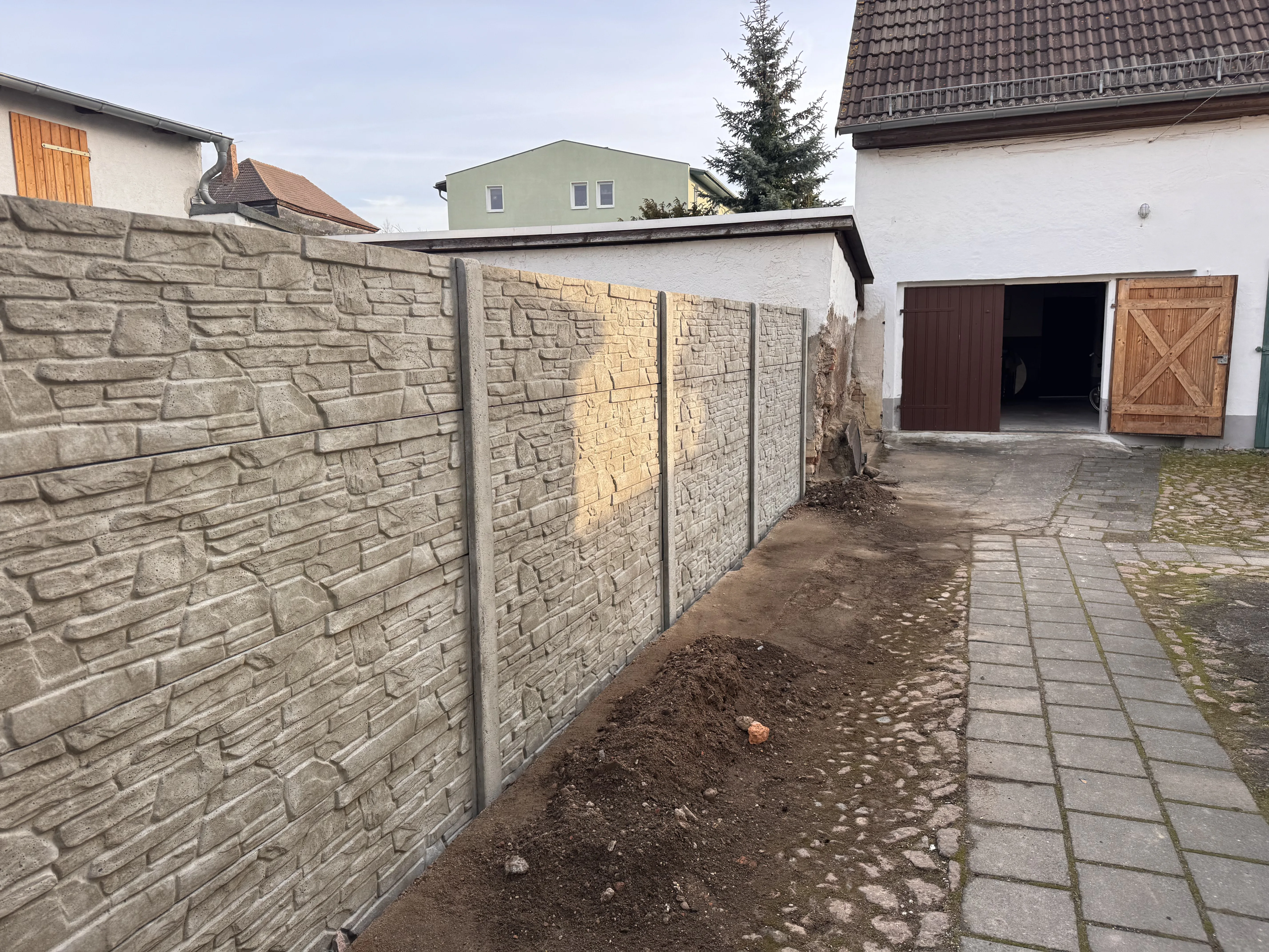 Moderner Betonzaun mit Steinoptik auf einem Grundstück, umgeben von unbefestigtem Boden